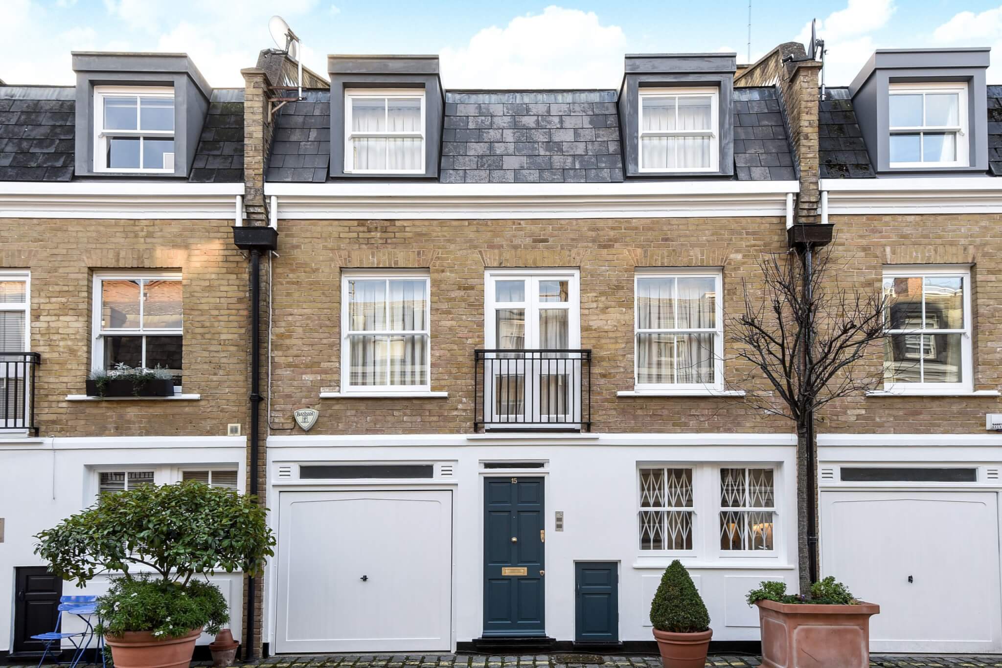 A row of two-story terraced houses with brick exteriors and dormer windows. Features include white window frames, a dark blue door, garages with white doors, and potted plants on a cobblestone street—perfect for those searching with Braithwait Estate Agents.