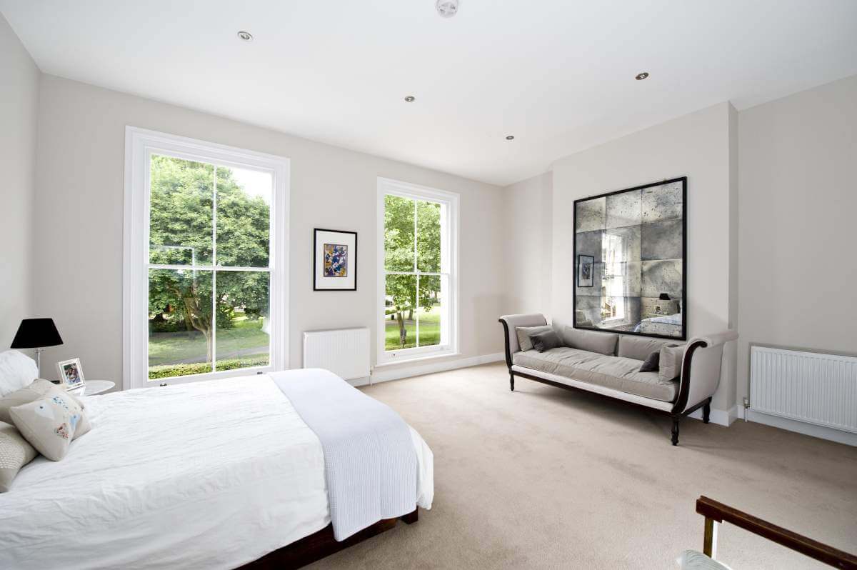 A bright, modern bedroom marketed by Braithwait Estate Agents boasts beige carpet and white walls. It features a bed with white bedding, a gray upholstered bench, and large windows revealing lush greenery outside. A decorative mirror and framed artwork add elegance to the space.