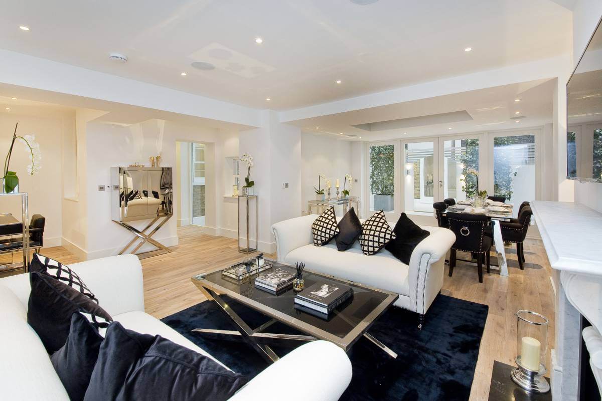 A modern living room curated by Braithwait Estate Agents showcases white sofas with black cushions, a glass coffee table atop a black rug, and a dining area featuring a sleek black table and chairs. Large windows and doors invite natural light, seamlessly connecting to the patio.