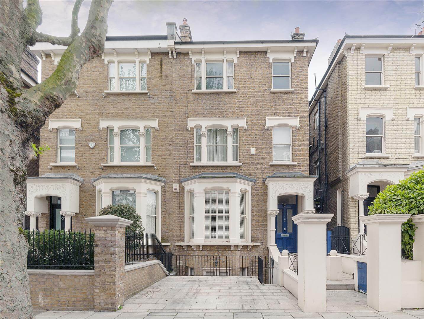 A row of three-story, brick Victorian-style houses in Queens Park, London, features ornate details, white trim, and multiple windows. A tree stands on the sidewalk in the foreground, accompanied by a low brick wall and iron fencing.