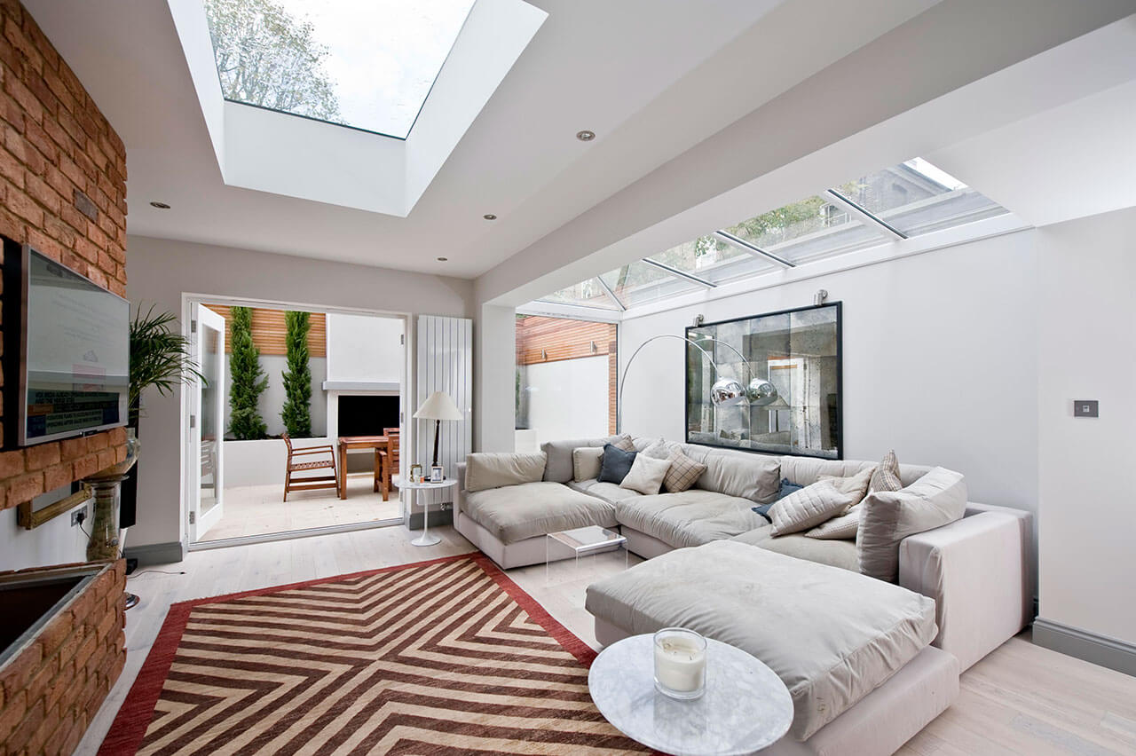 A modern living room curated by Braithwait Estate Agents features a skylight, large sectional sofa, and zigzag patterned rug. The space leads to a room with a fireplace. A marble side table and tall plants grace the window, while walls and decor are in soothing neutral tones.