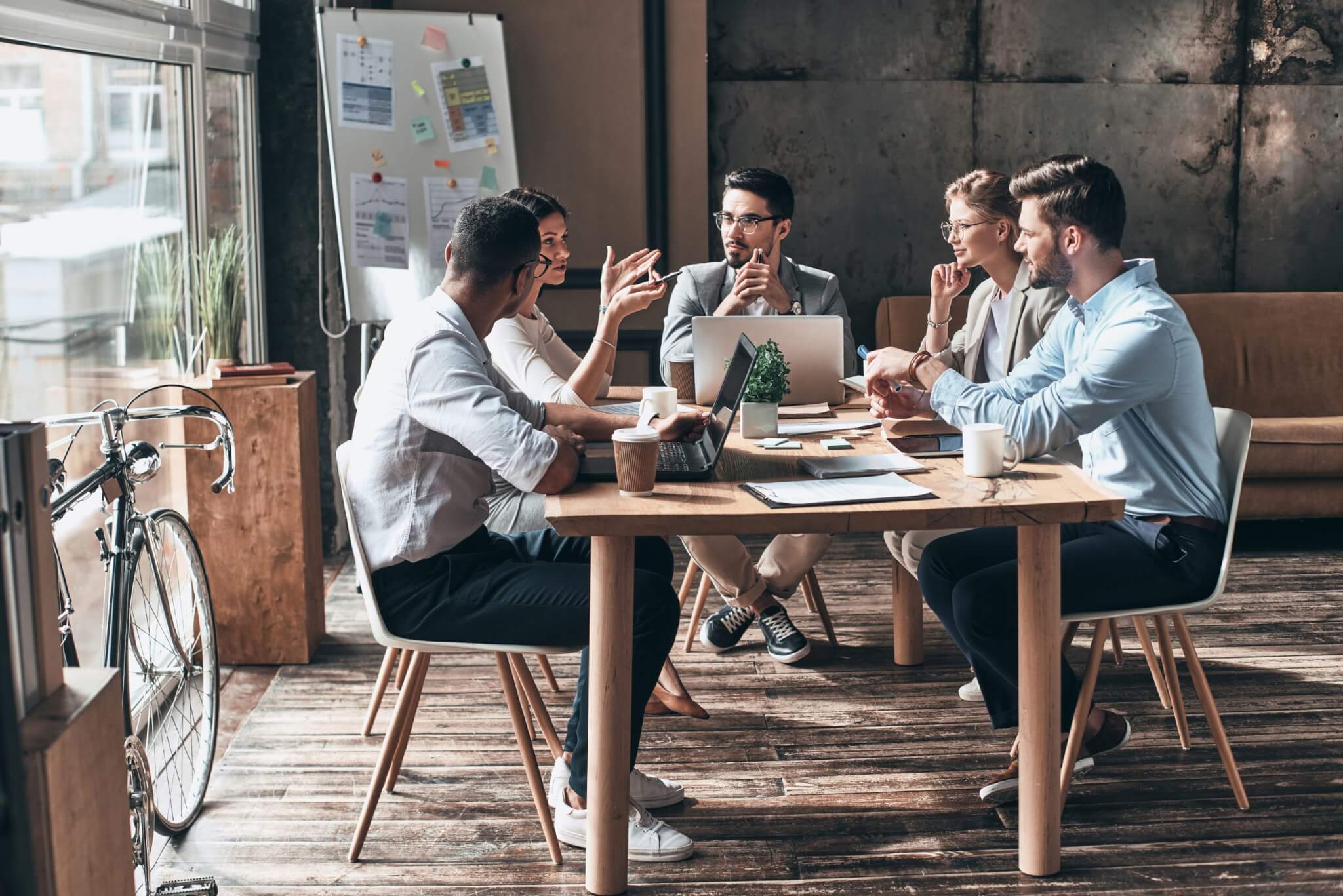 Five people are gathered around a wooden table in Braithwait Estate Agents' modern office. They're deep in discussion, surrounded by laptops, notebooks, and coffee cups. In the background, a bicycle leans against the wall beside a whiteboard filled with notes.