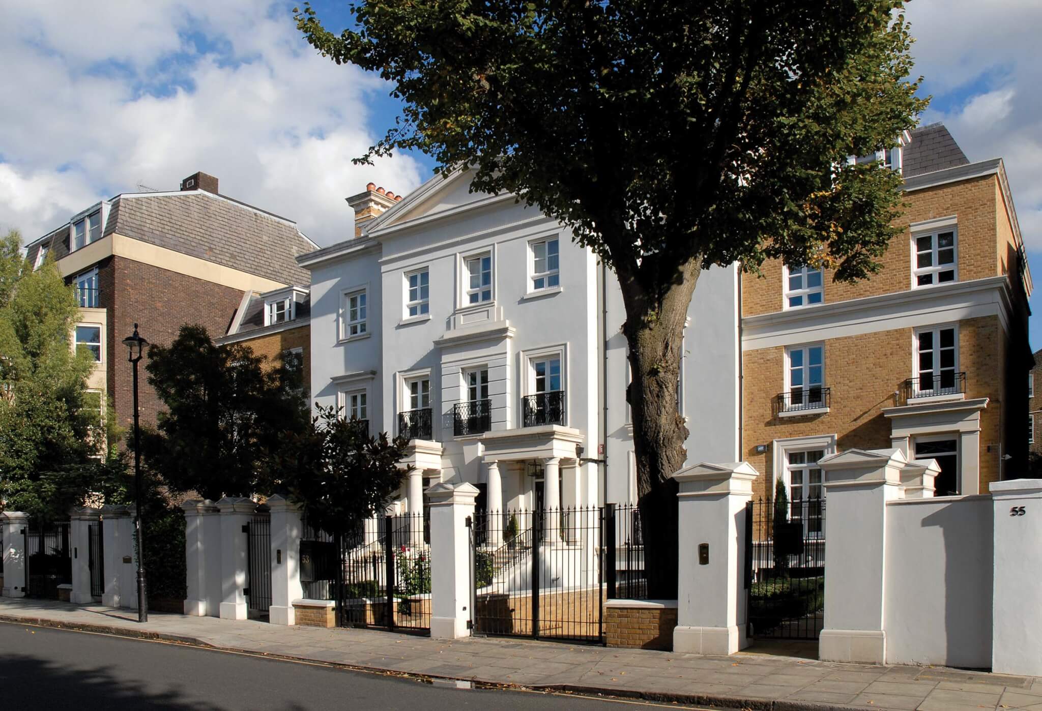 A row of elegant townhouses lined along a quiet street in St John’s Wood, London. The white and brick exteriors are complemented by black iron gates and mature trees, all under a partly cloudy sky. The architecture is classic and well-maintained.