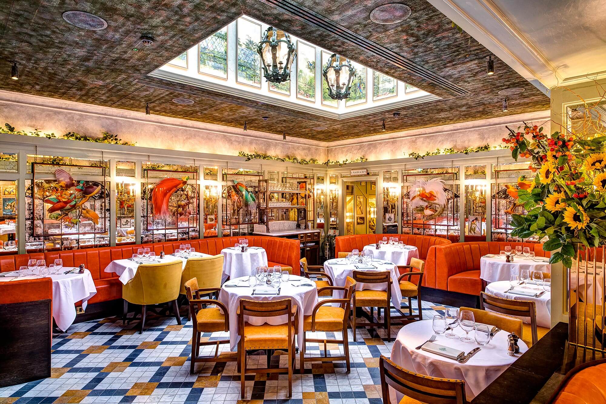 A vibrant, upscale restaurant in St John’s Wood, London, boasts an interior with round tables draped in white tablecloths, colorful chairs, and decorative stained glass. A skylight illuminates the floral arrangements while mirrored walls and intricate artwork enhance the elegant atmosphere.