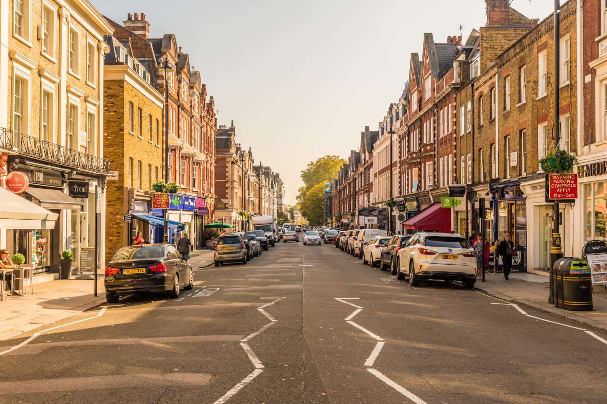 A sunny street in St John’s Wood, London, is lined with brick buildings and shops on both sides. Parked cars occupy the road while a few pedestrians stroll along the sidewalks. The clear sky and long shadows from the buildings hint at a late afternoon setting.