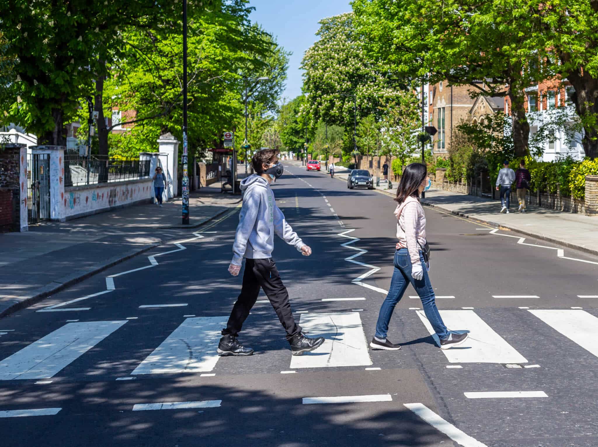 Two people stroll along a zebra crossing on a tree-lined street in St John’s Wood, London. Its a sunny day, and they head in the same direction on a mostly empty road, with only a few others visible in the distance.
